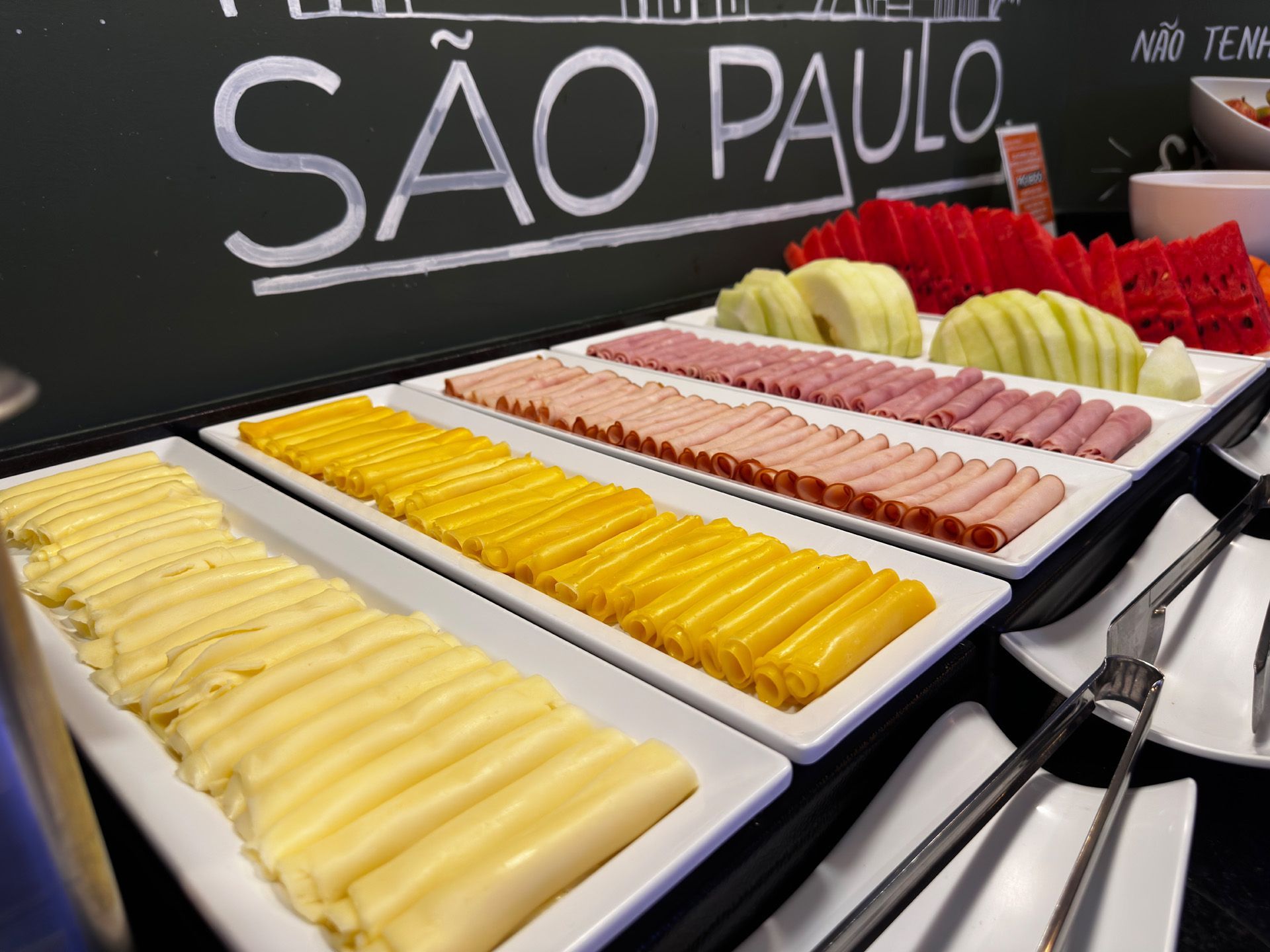 Buffet com exposição de queijos fatiados, frios e frutas em São Paulo, Brasil.