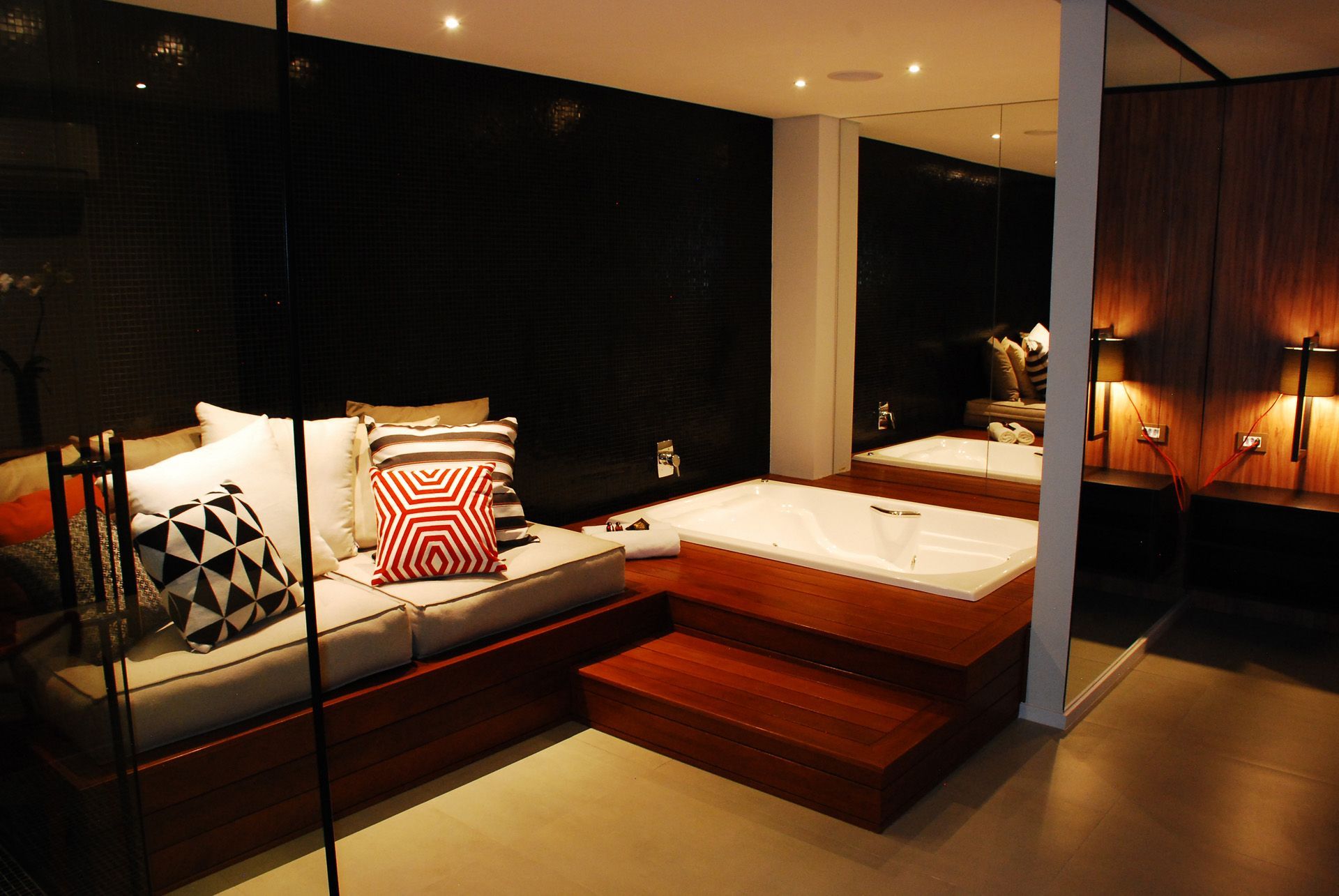 Quarto aconchegante com banheira de hidromassagem elevada e sofá com almofadas decorativas. Paredes escuras, espelhos e iluminação quente.