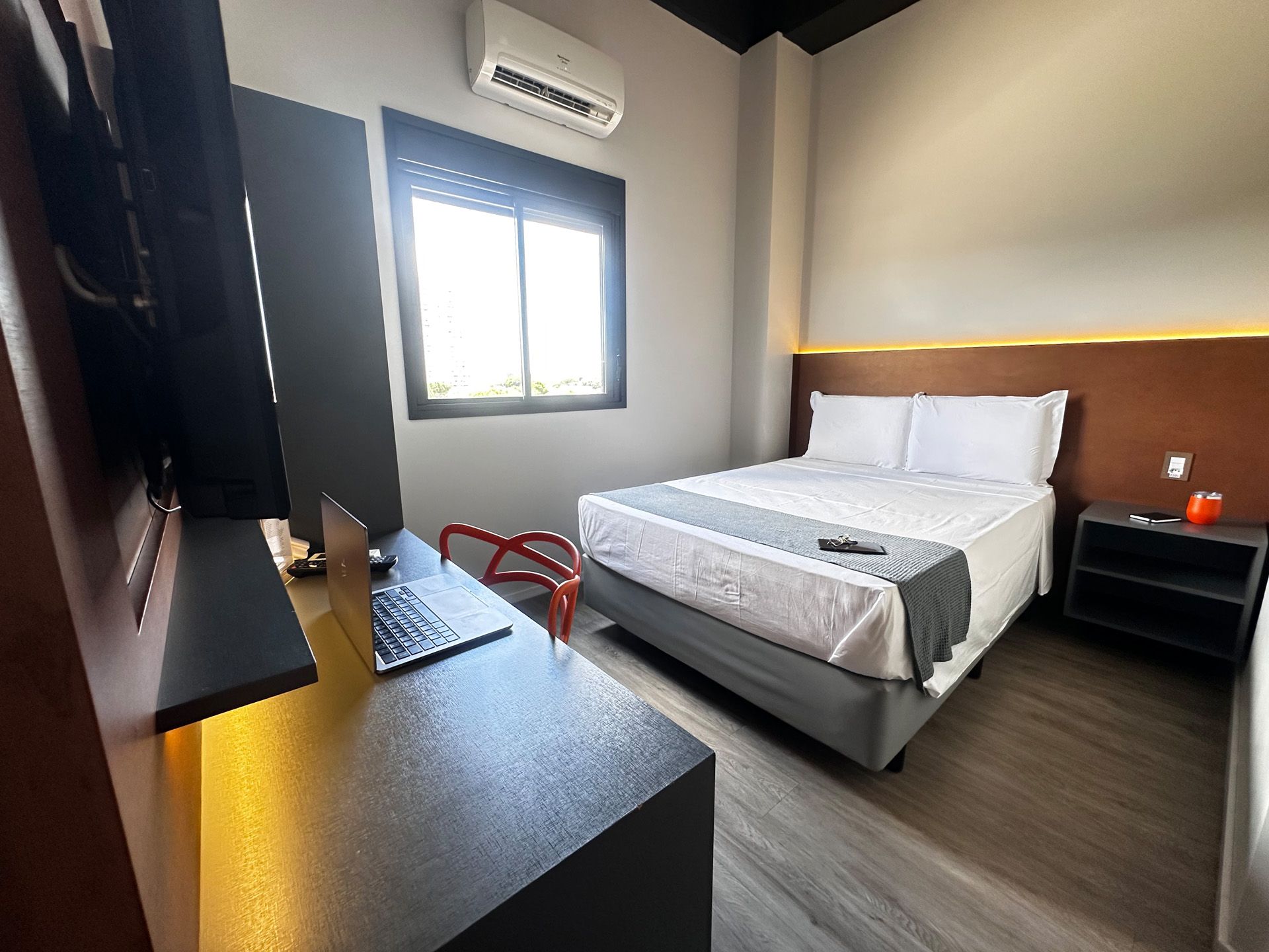 Quarto de hotel com cama, mesa com laptop, janela e ar condicionado.