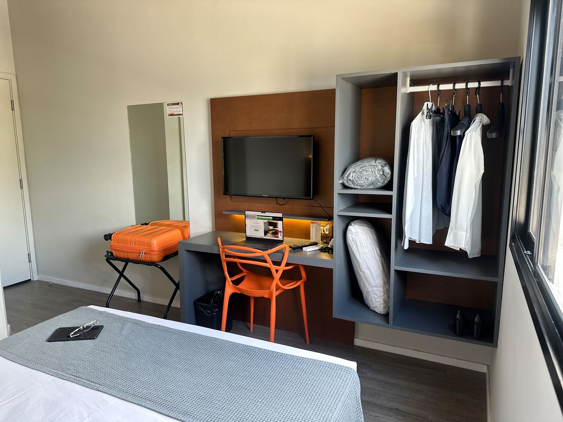 Interior de quarto de hotel com cama, escrivaninha, guarda-roupa, cadeira laranja, bagagem e TV.