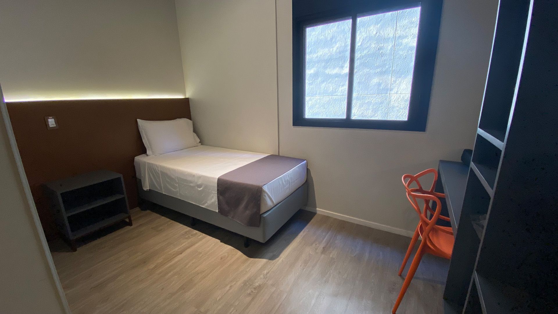 Quarto pequeno com cama de solteiro, mesa de cabeceira, escrivaninha e janela. As paredes são neutras, com detalhes em madeira.