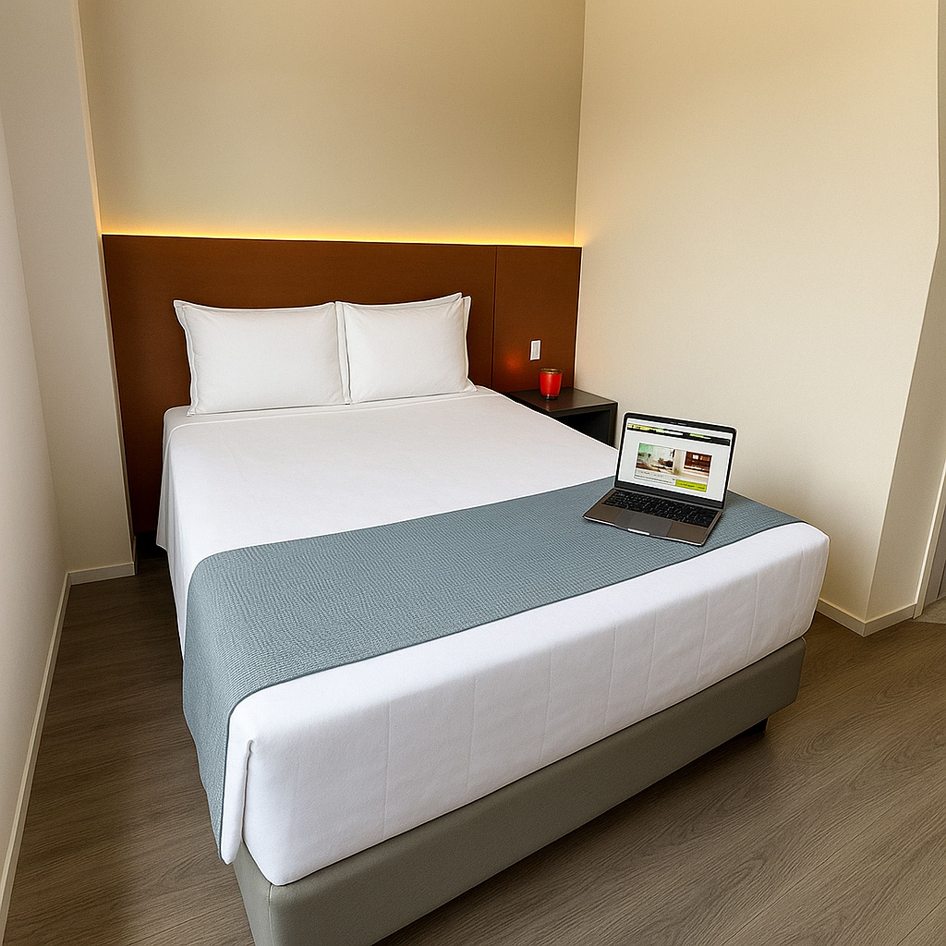 Um quarto de hotel com cama, laptop e mesa de cabeceira. Paredes bege, cama cinza, cabeceira marrom.