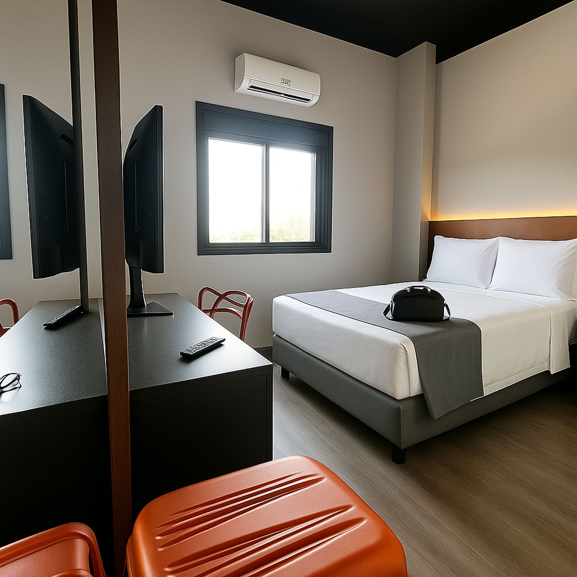 Quarto de hotel: cama, escrivaninha com dois monitores, janela aberta, malas laranja.