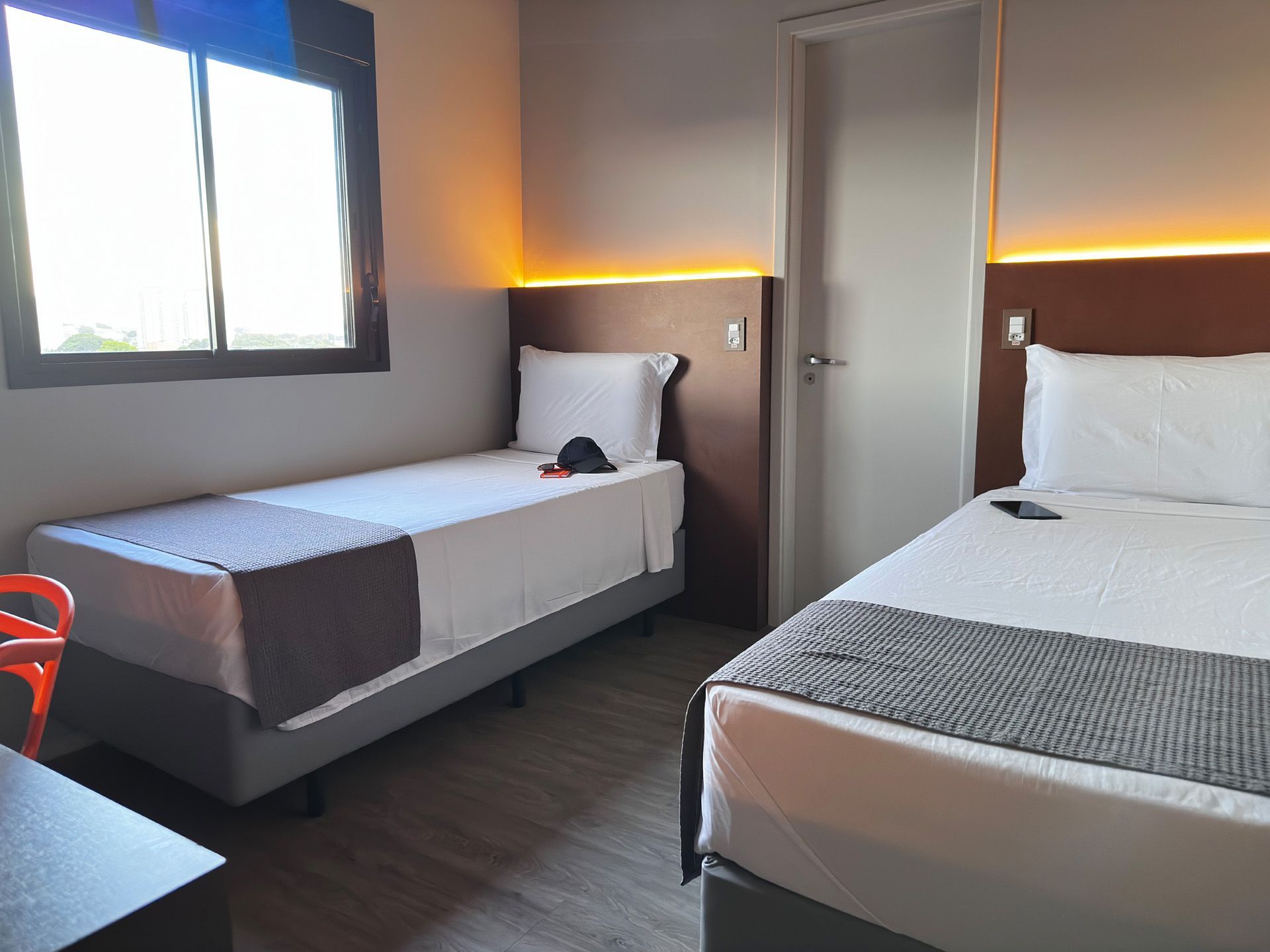 Quarto de hotel com duas camas de solteiro, roupa de cama cinza e branca, janela pequena e cadeira de escritório laranja.