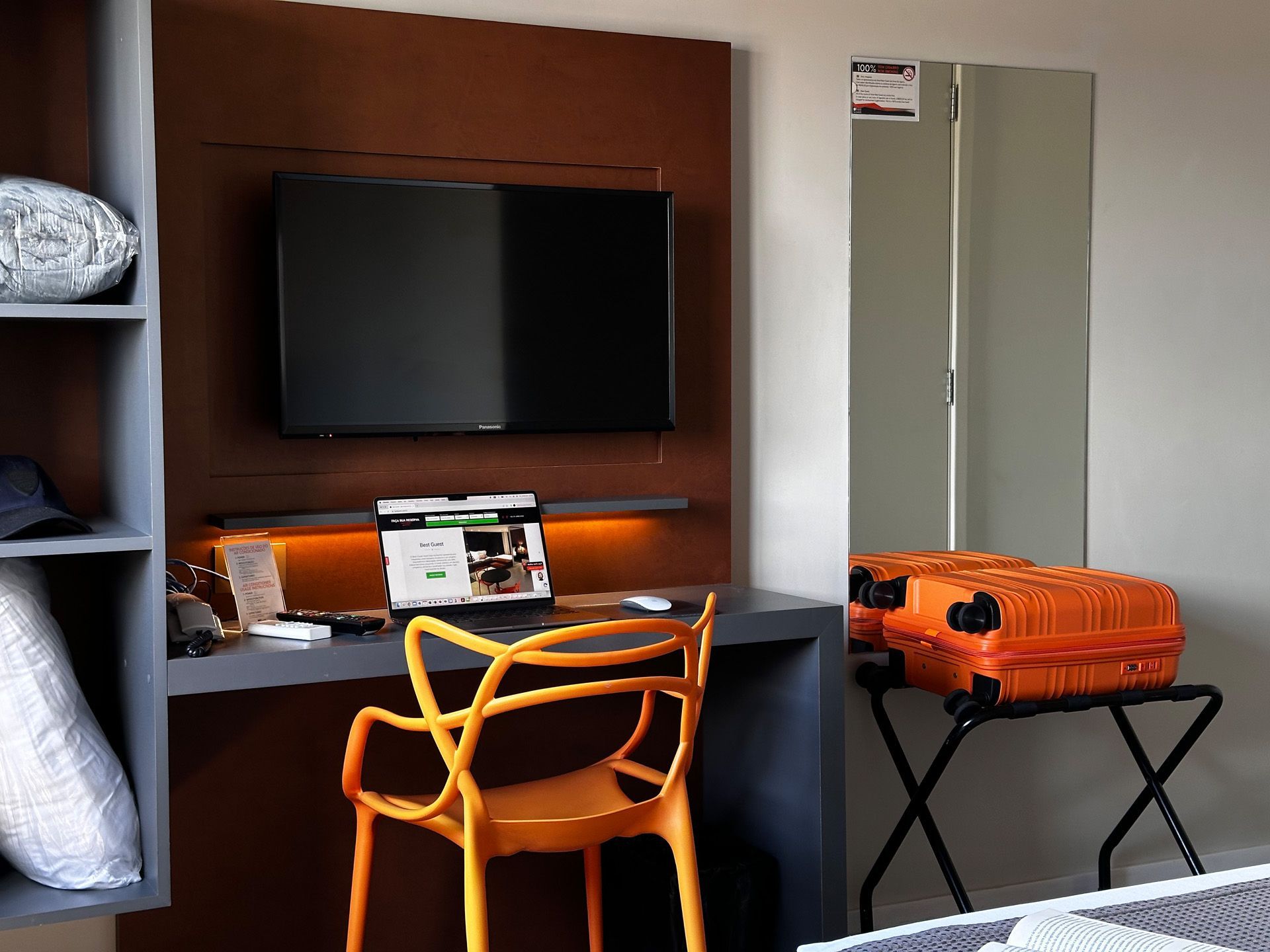 Interior de quarto de hotel com escrivaninha, TV, cadeira laranja, bagagem e prateleiras embutidas.