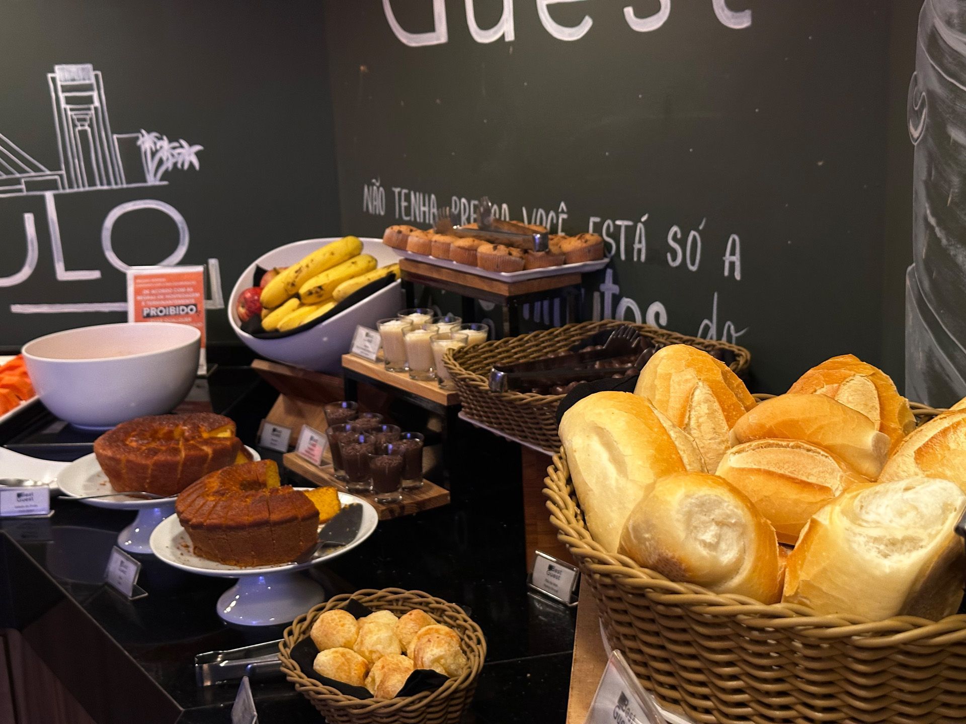 Buffet de café da manhã: Pães, doces, frutas e queijos em um balcão preto, ao lado de uma parede de lousa.