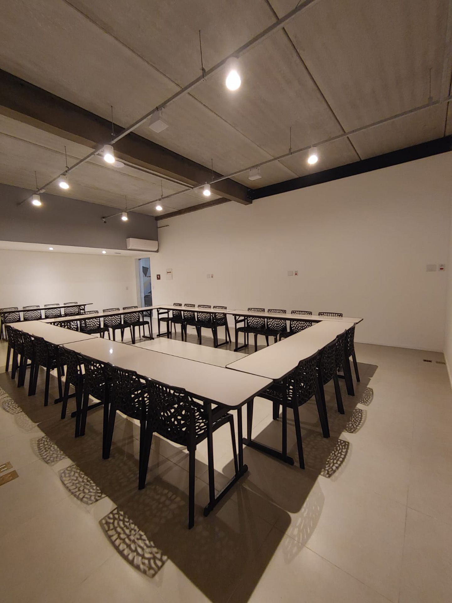 Sala de reuniões com mesas quadradas e cadeiras pretas, sob um teto de concreto com iluminação.