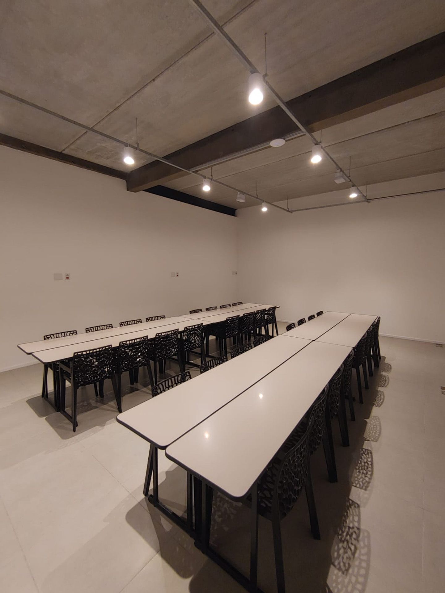 Sala de conferências vazia com mesas compridas, cadeiras pretas e iluminação suspensa. Paredes brancas, piso cinza.
