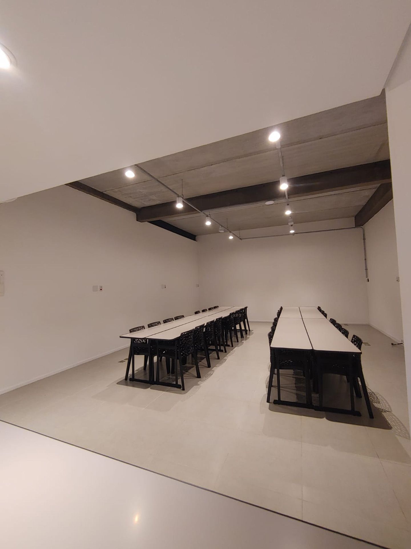 Sala moderna e vazia com duas fileiras de mesas e cadeiras, sob um teto de concreto com luzes.