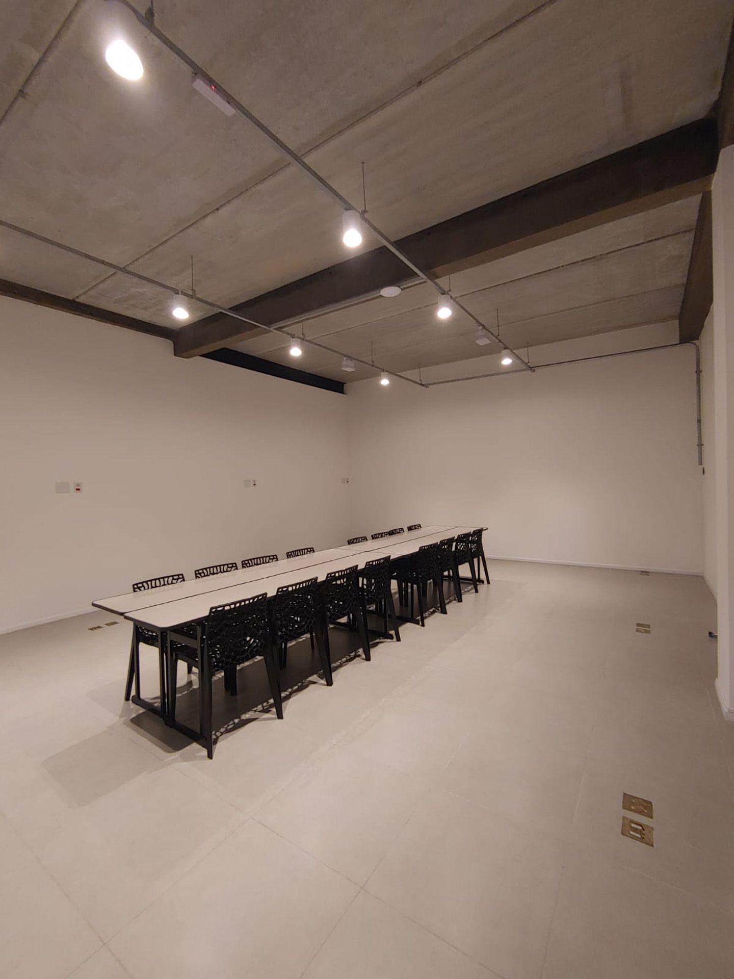 Sala de conferências vazia e bem iluminada, com uma longa mesa e cadeiras. Paredes bege, teto de concreto e piso de azulejo.