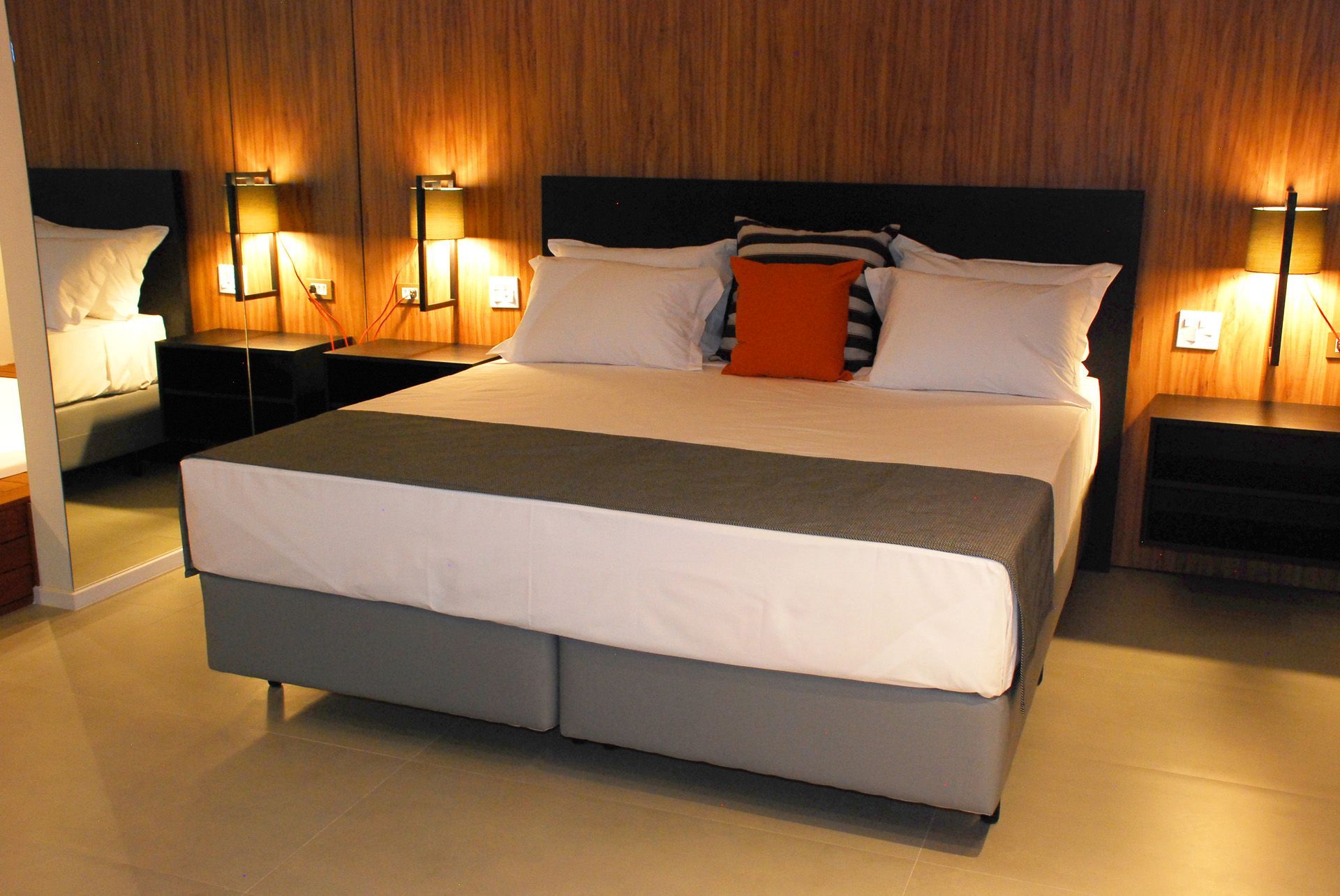 Quarto com uma cama grande, mesas de cabeceira e apliques de parede, refletidos em um espelho.