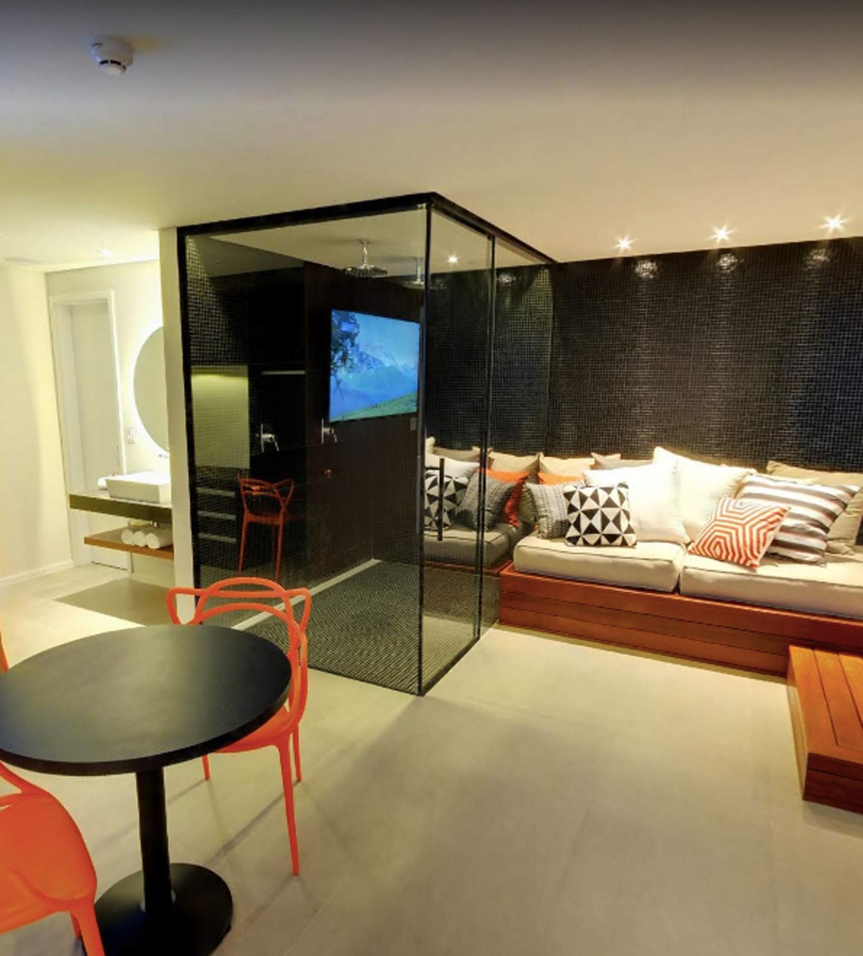 Interior de suíte de hotel moderna com área envidraçada emoldurada em preto, contendo TV, escrivaninha e banheiro. Um sofá e uma mesa estão próximos.