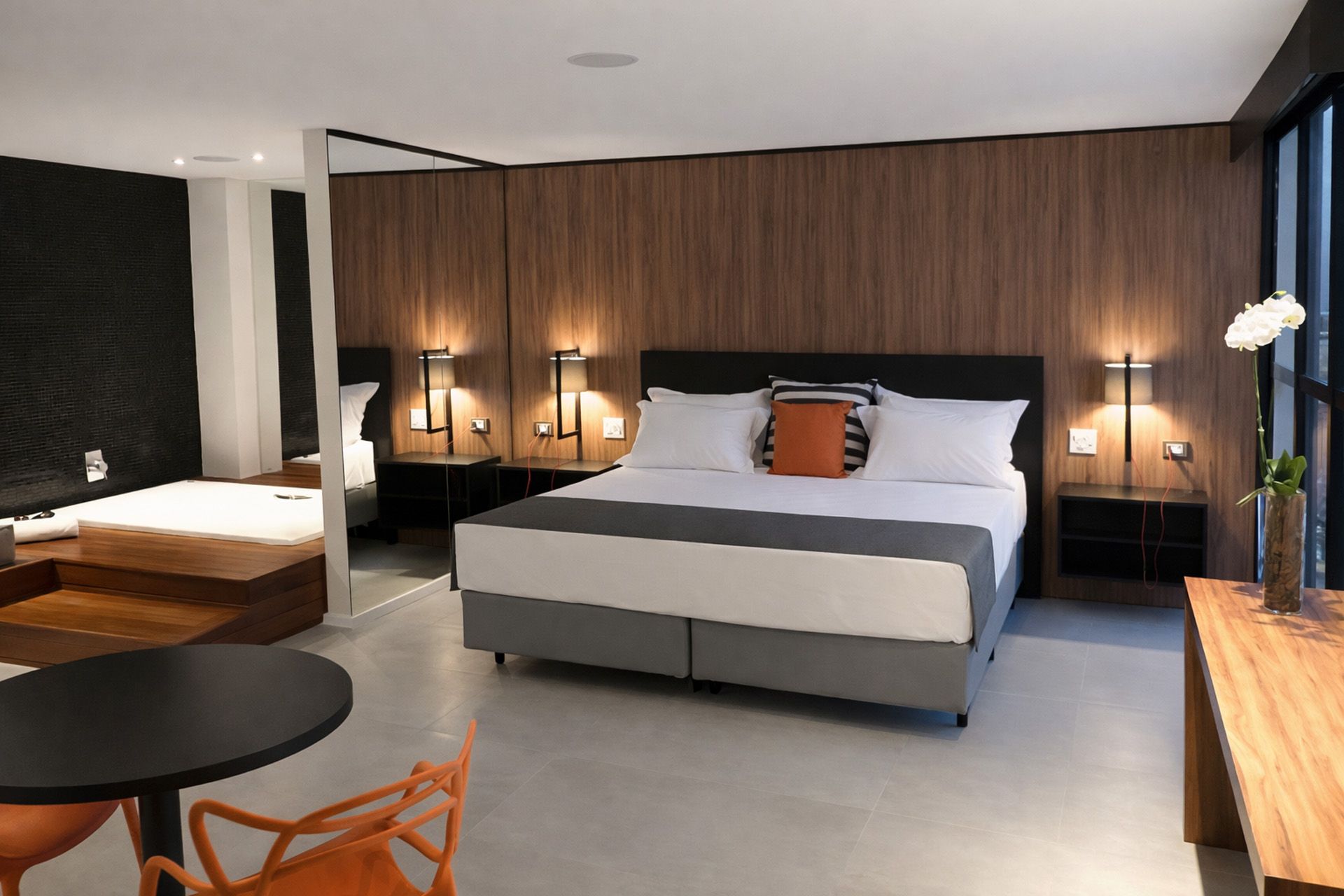 Quarto de hotel com cama king-size, jacuzzi, mesa e cadeiras laranja. Painéis de madeira escura e iluminação diferenciada.