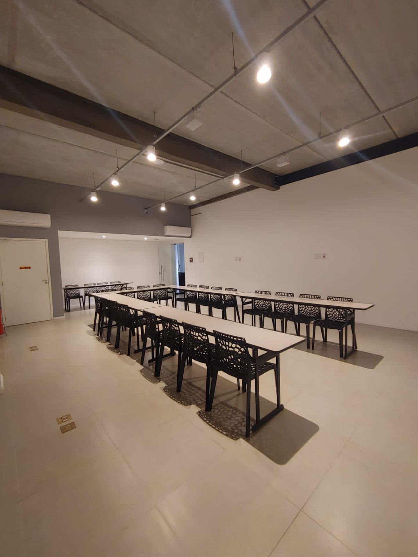 Sala de conferências vazia com mesas compridas e cadeiras pretas, sob luzes de teto brilhantes.