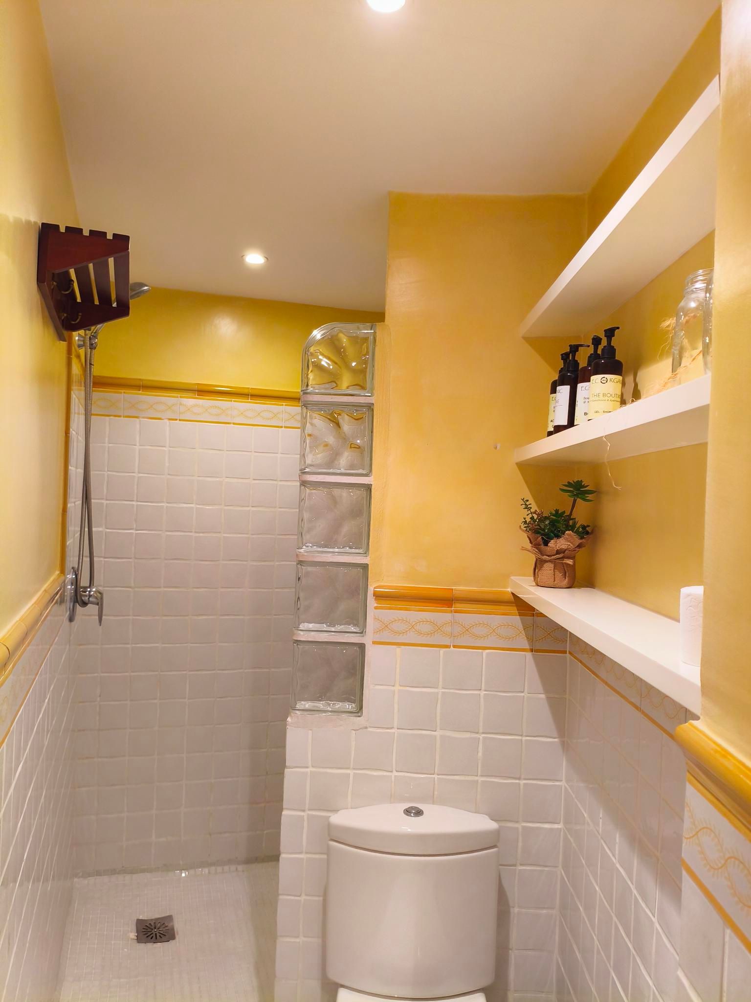 Yellow bathroom with a shower, toilet, and shelves. Glass blocks and white tile.