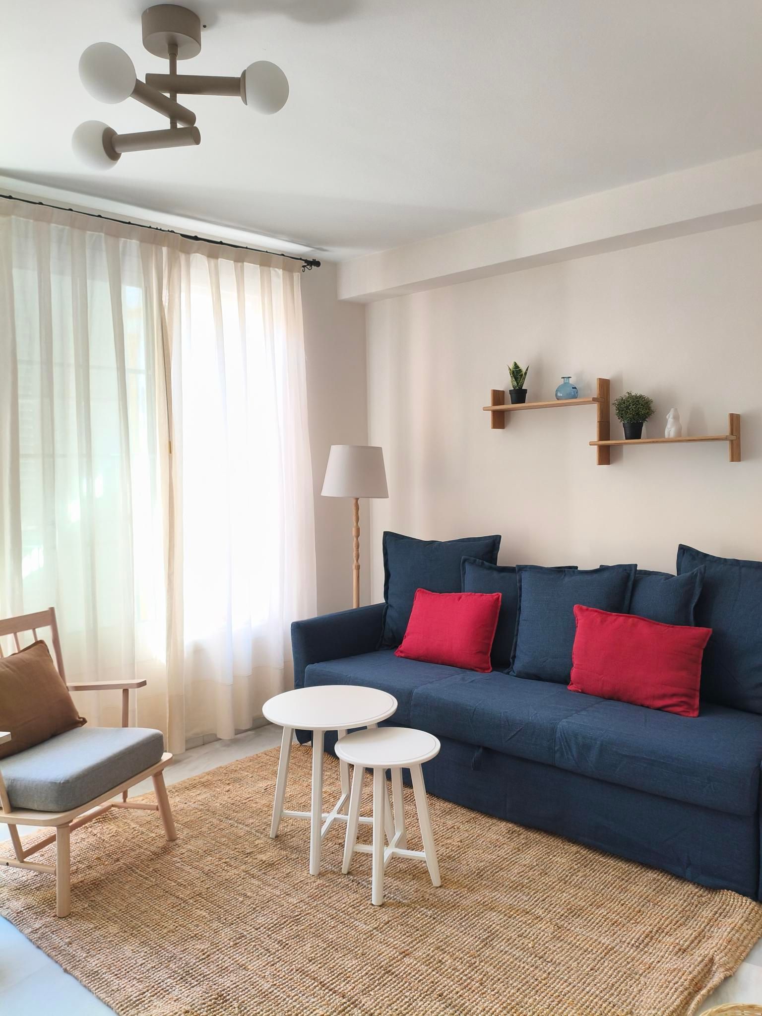 Cozy living room with blue sofa, red pillows, natural rug, small white tables, and wooden shelves.