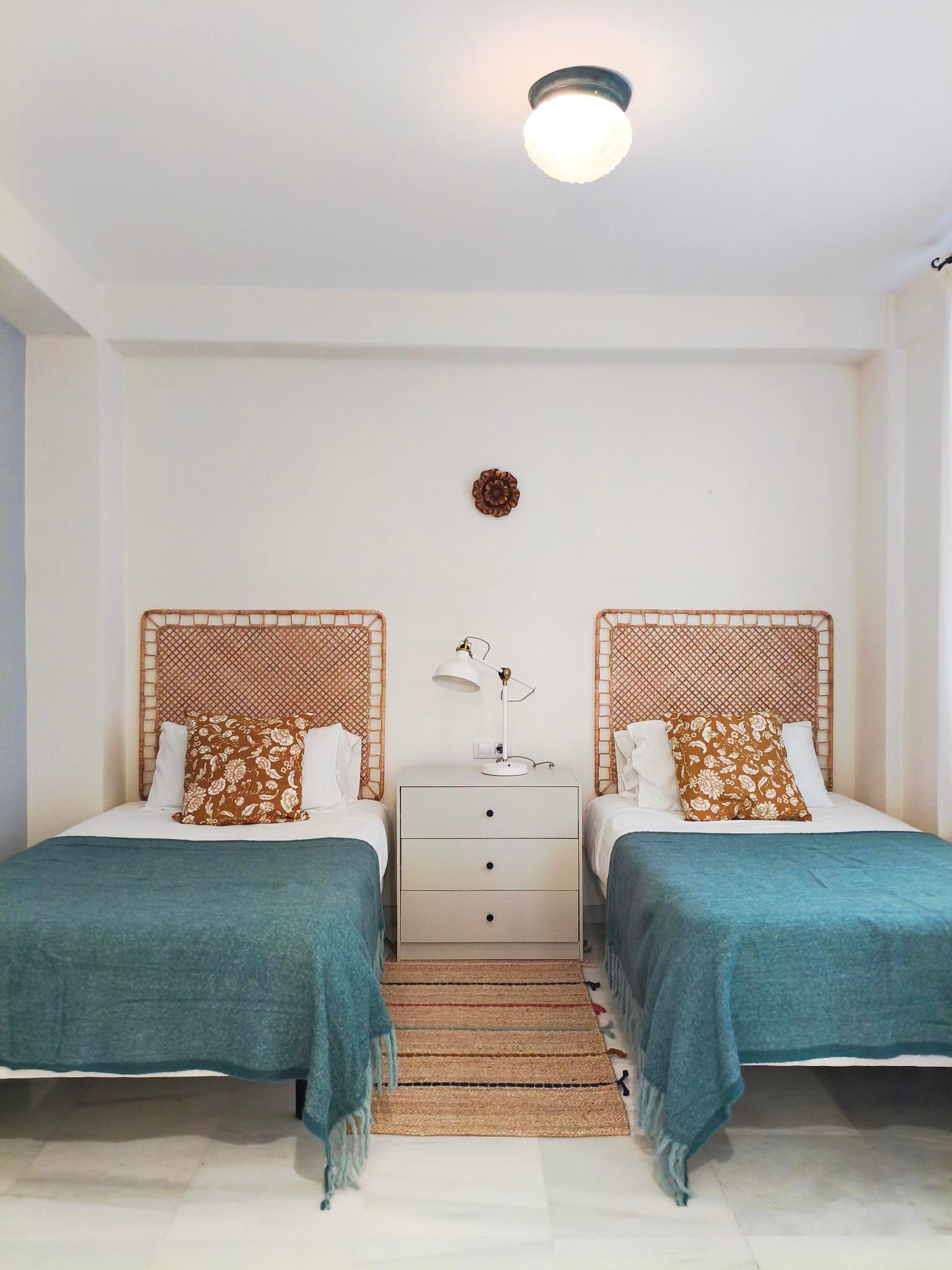 Two twin beds with teal blankets and woven headboards in a bright room. A nightstand and a small rug are between the beds.