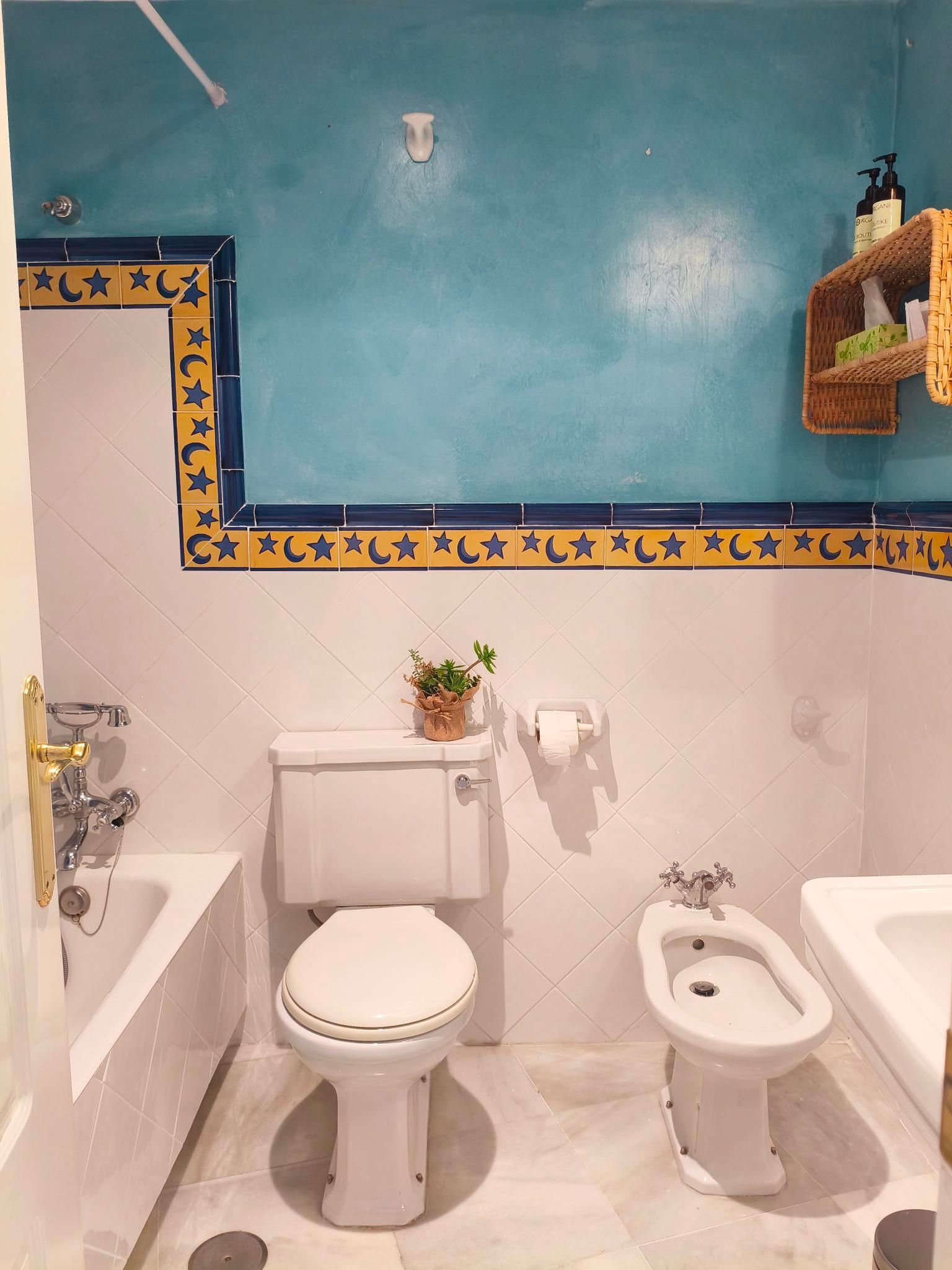 Bathroom with blue wall, white tiles, toilet, bidet, tub, gold fixtures, and a wooden shelf.