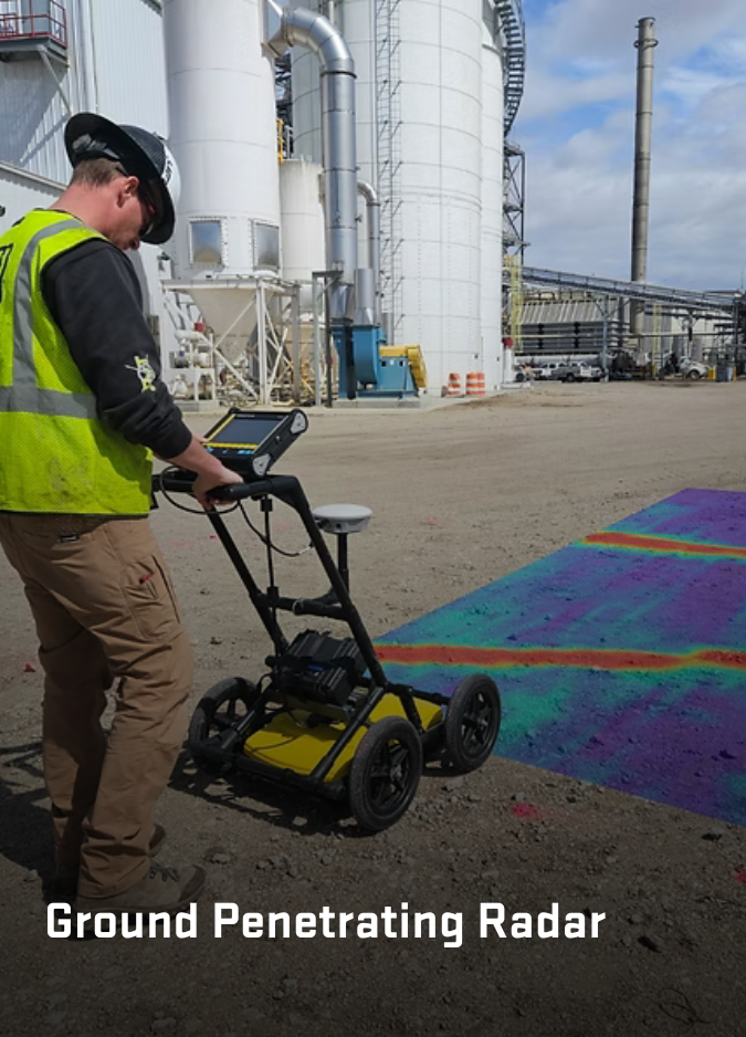 A worker in a high-visibility vest uses Ground Penetrating Radar to scan a gravel area near an industrial facility.