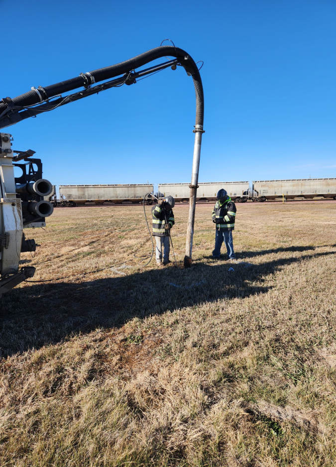 Two workers in high-visibility gear use a vacuum truck boom to excavate a hole in a dry, open field near a train.