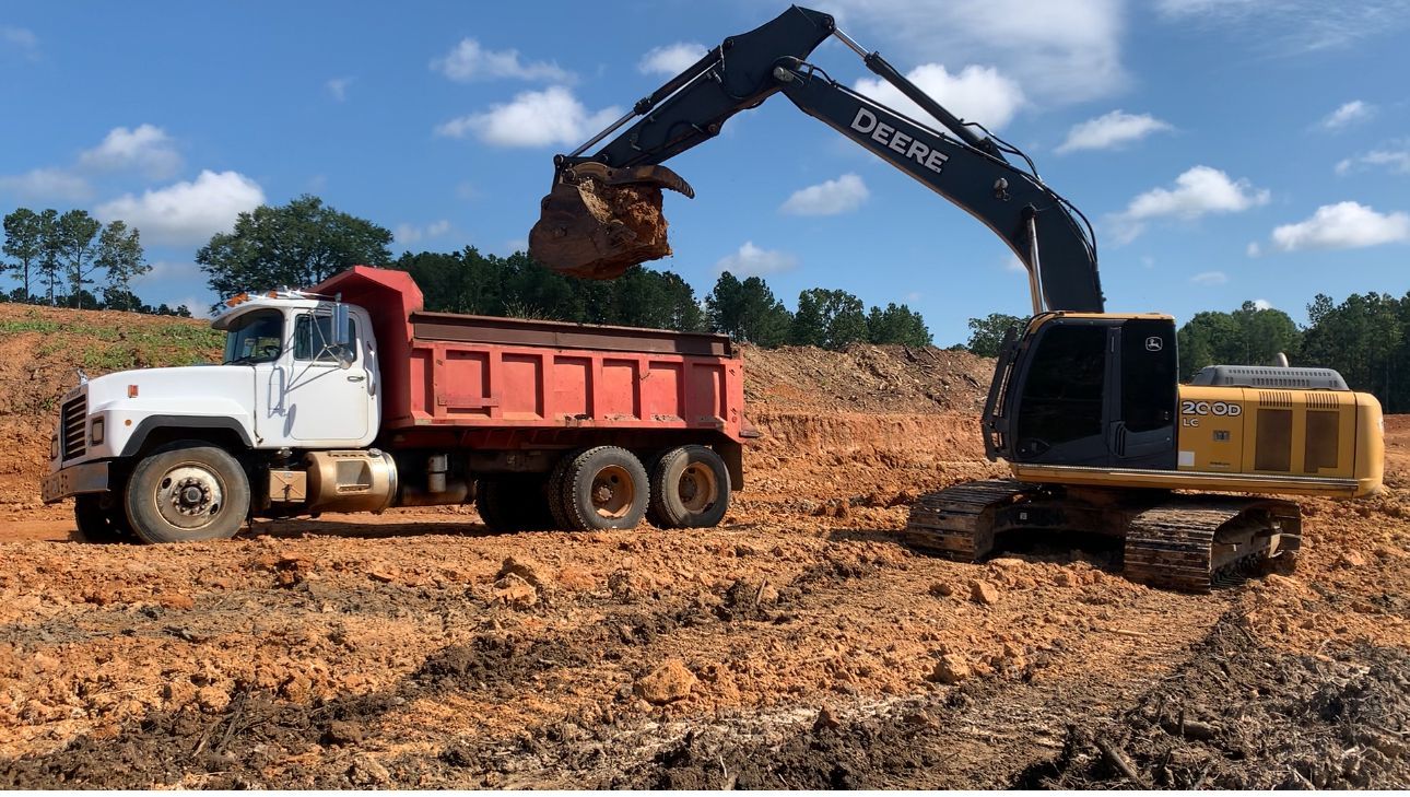 A John Deere excavator loading red-dump truck with dirt at a construction site, blue sky background.