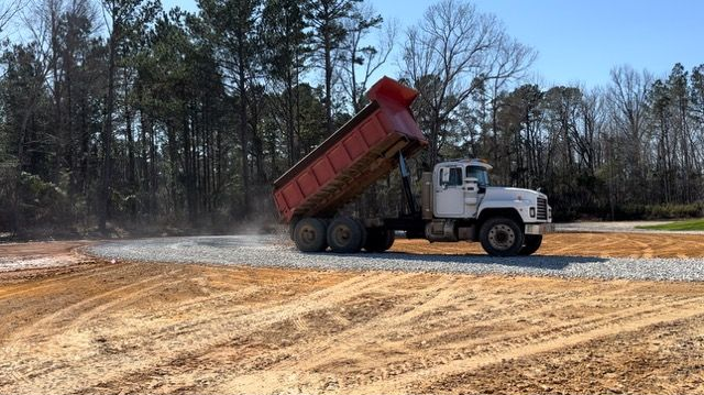 A dump truck unloading gravel on a dirt road, with trees and blue sky in the background.