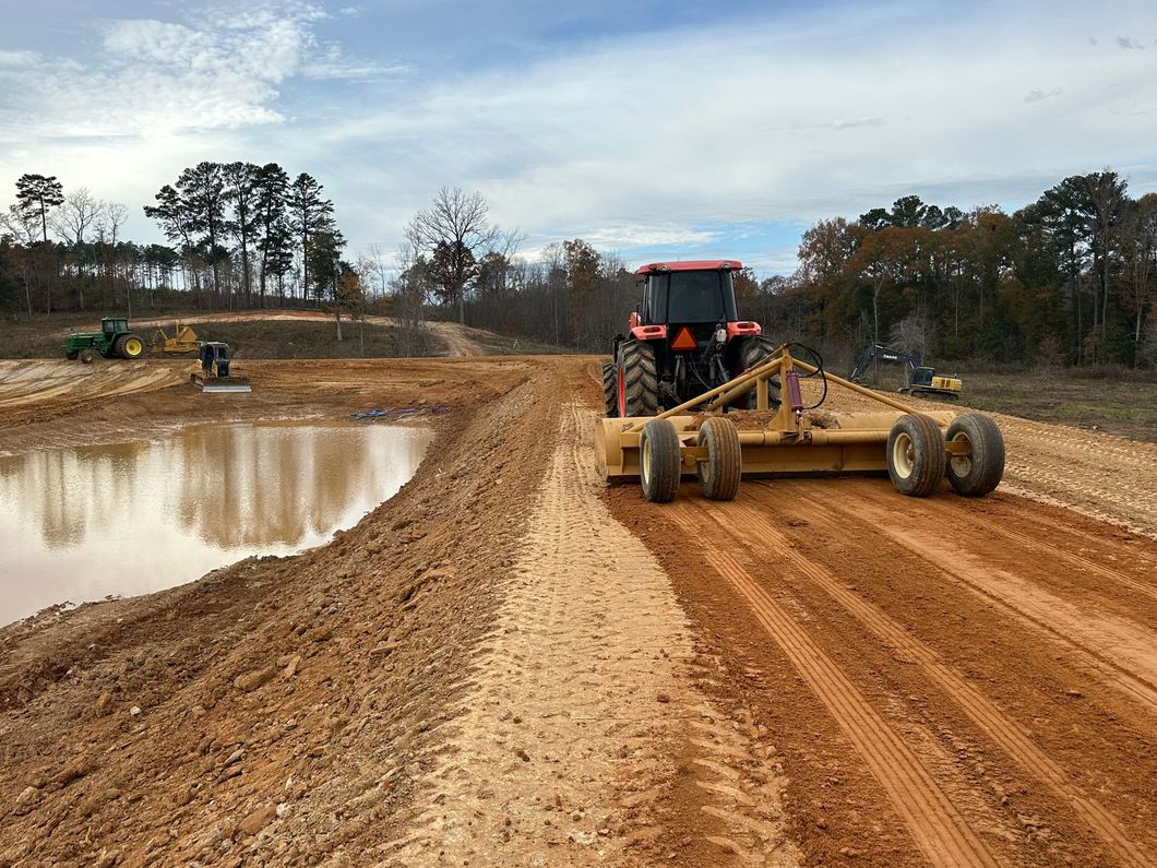 Red tractor pulling a road grader levels a gravel road next to a pond on a construction site.