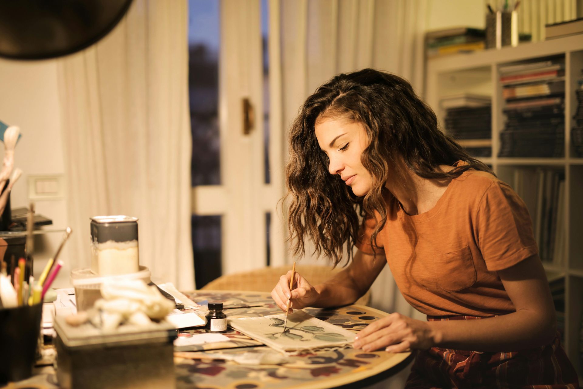 Woman with curly hair painting at a table in a well-lit room.