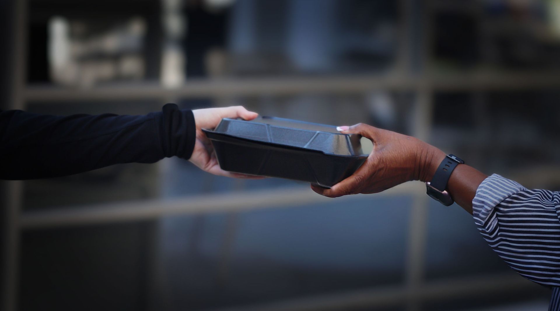 Hands passing a black food container outside a building.