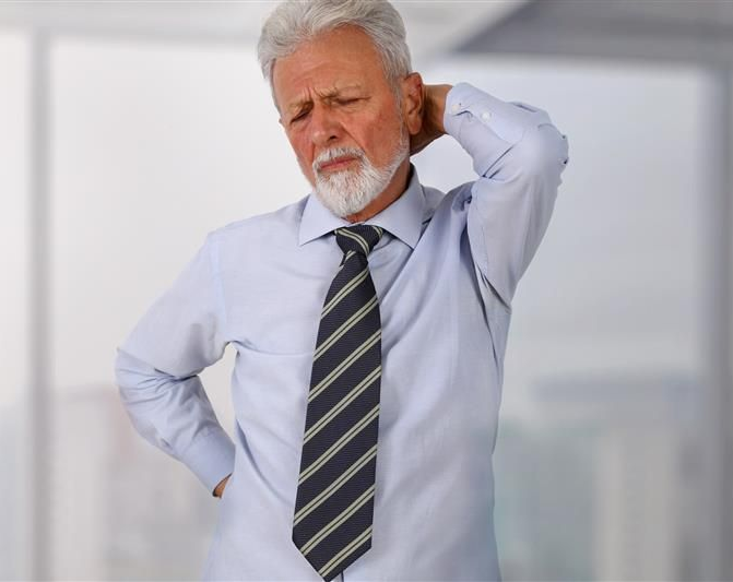 Man in office setting, holding his back and neck, showing signs of pain.