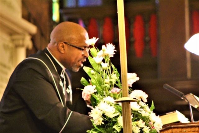 Rev. Manning in a black robe by arranged flowers near a lit candle, inside a church.