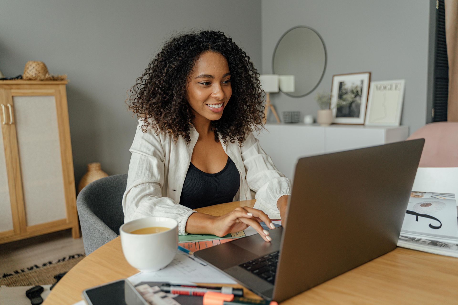 Woman with curly hair smiles while working on a laptop at a round table, coffee nearby.