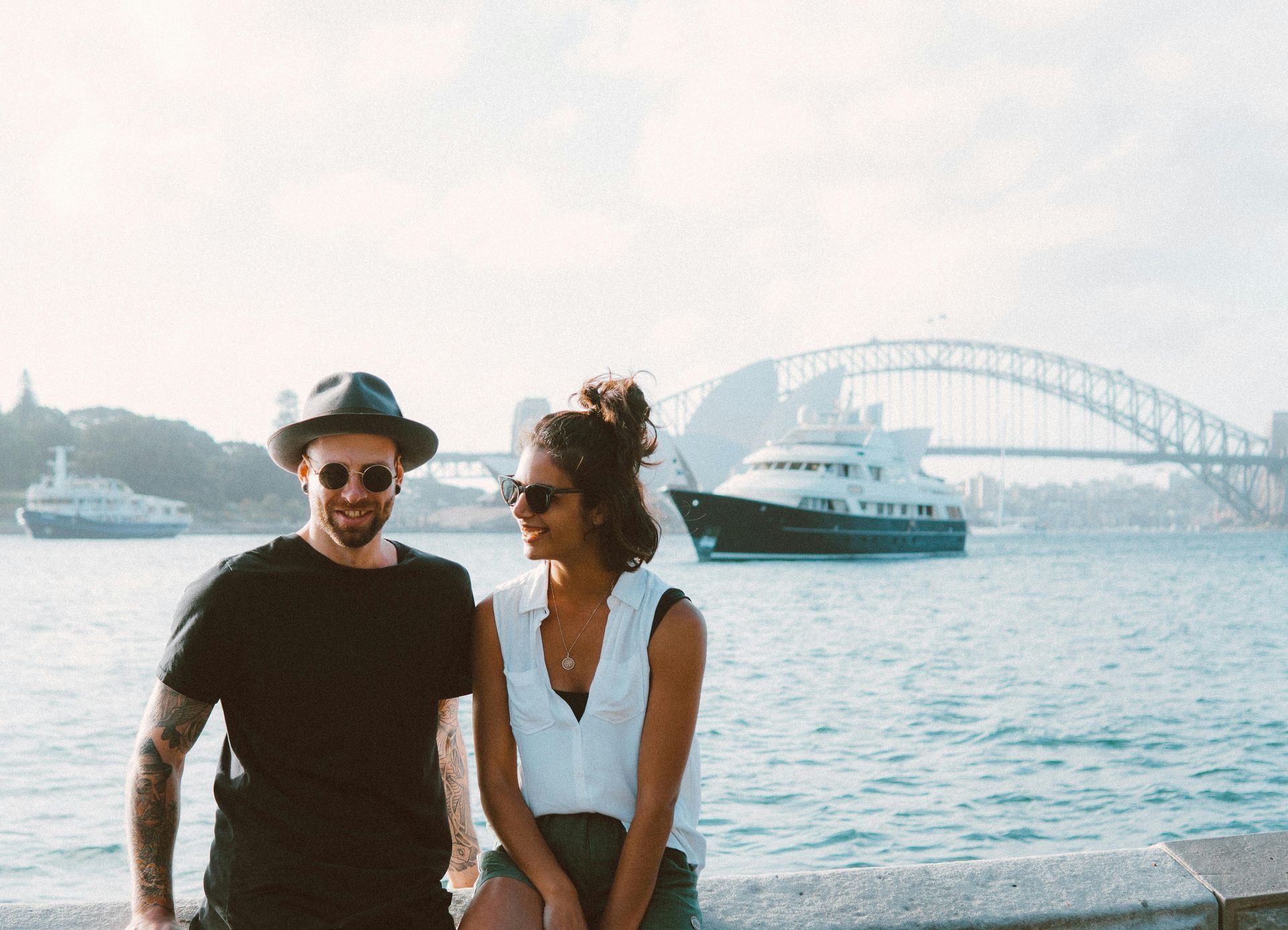 Couple smiling near water with the Sydney Harbour Bridge and a boat in the background. Sunny day.