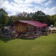 Rustic cabin with red roof and stone foundation on a grassy lawn, surrounded by trees under a cloudy sky