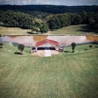 Rural lakeside pavilion with red roof on a grassy lawn, backed by trees and hills