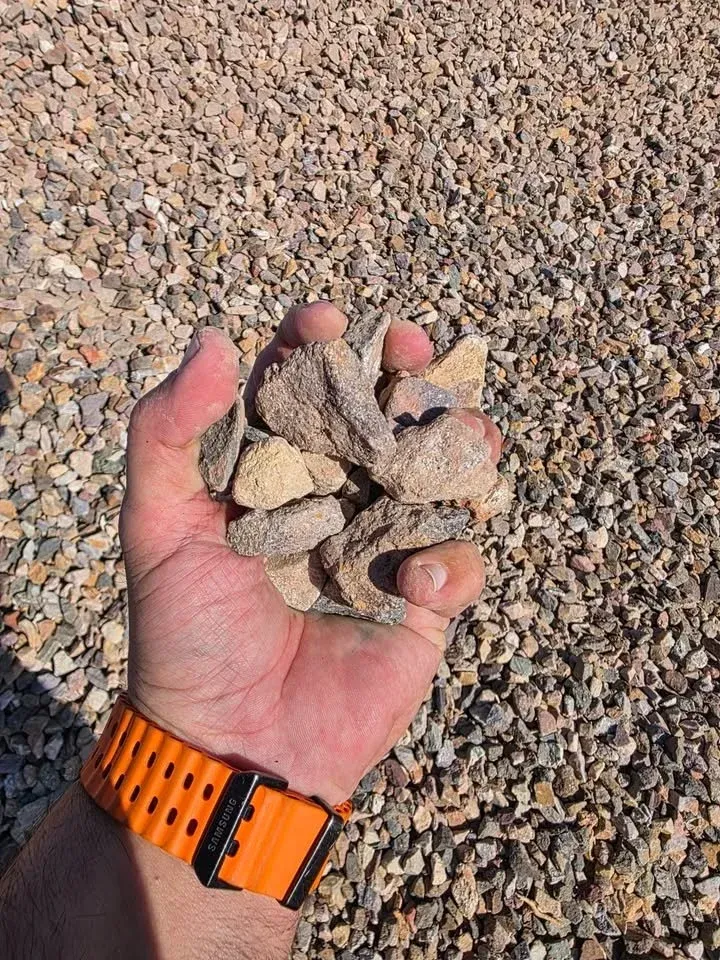 Hand holding a pile of gravel, with more gravel in the background. An orange watch is visible on the wrist.