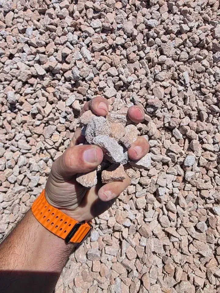 Hand holding a pile of light-colored gravel against a larger bed of similar gravel; orange watch on wrist.