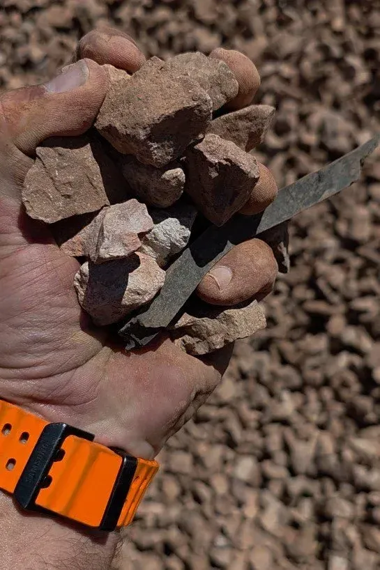 Handful of reddish-brown rocks and a gray piece held by a person with an orange watch in a rocky outdoor setting.