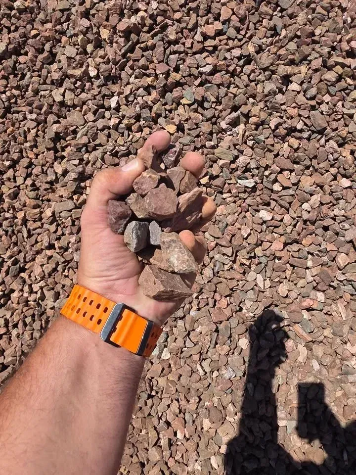 Handful of brown rocks, held over a pile of similar rocks. Orange watch on wrist. Sunny outdoor setting.