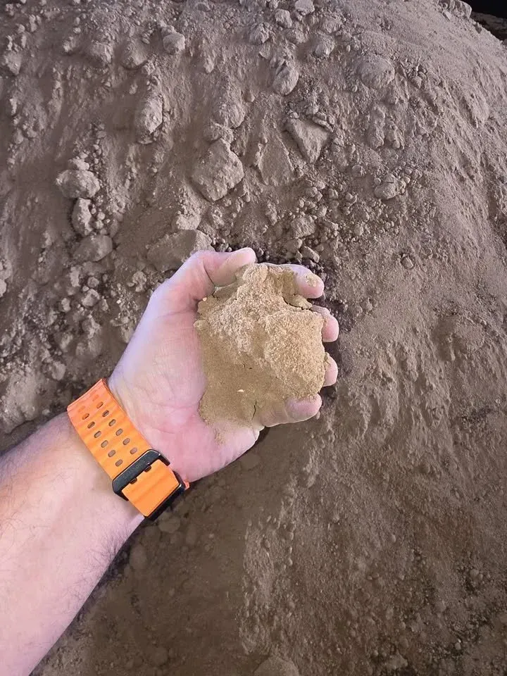 Hand holding a clump of light brown soil in front of a larger pile of the same soil.