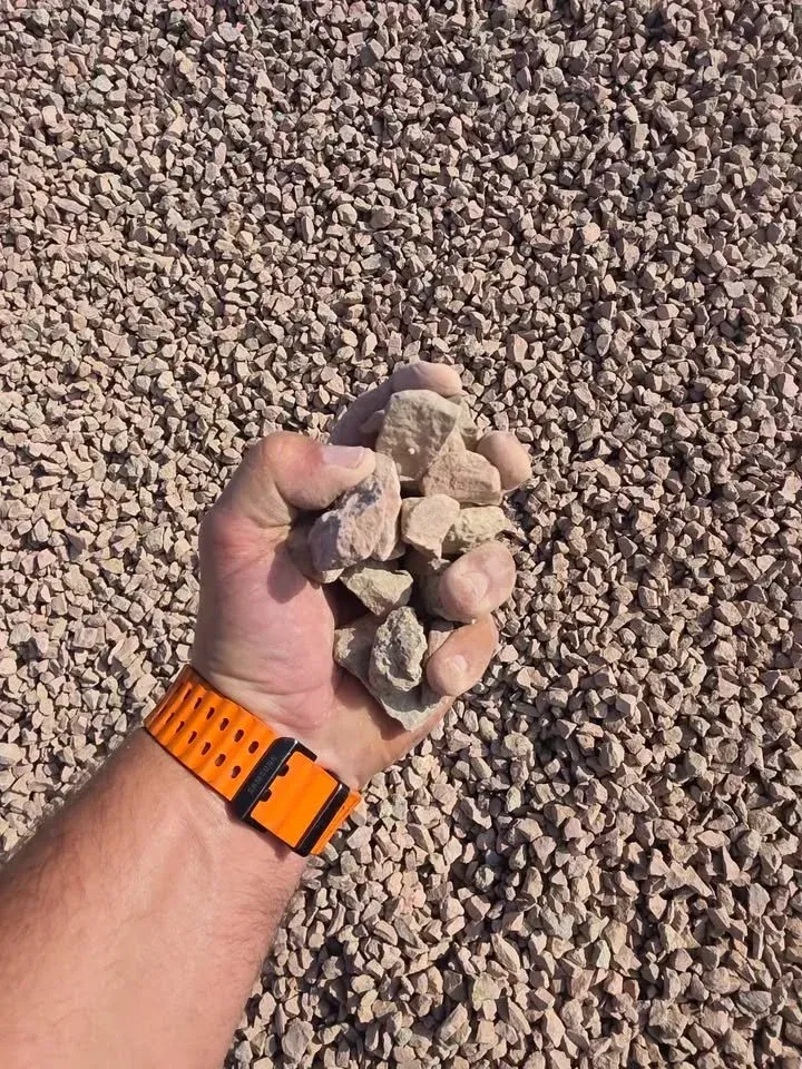 Handful of beige gravel, held against a backdrop of more gravel. Orange watch band visible.