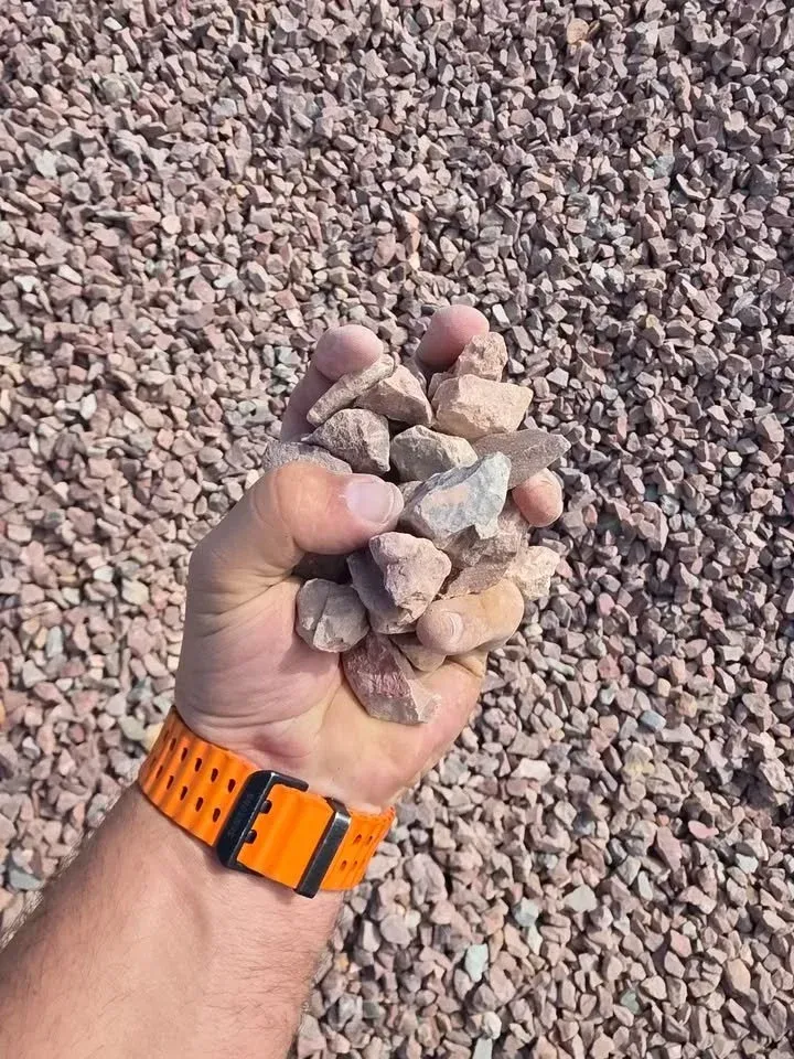 Handful of light-colored gravel against a larger gravel bed. Orange watch band visible on wrist.