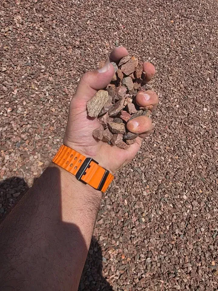 Person's hand holding a pile of reddish-brown gravel, wearing an orange watch. Set on a gravel ground.