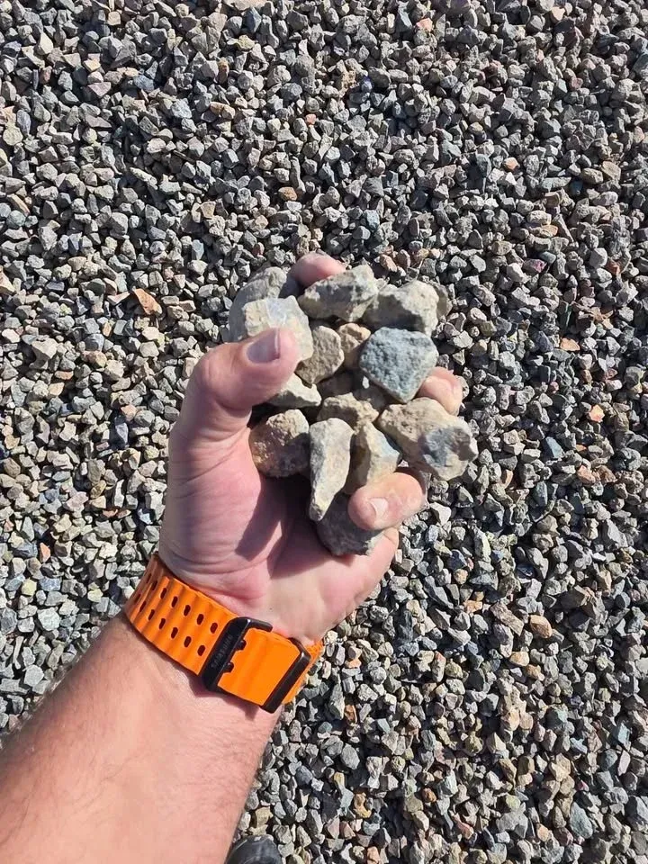 Hand holding a pile of gray and tan gravel. Bright orange watch on wrist. Ground covered in gravel.