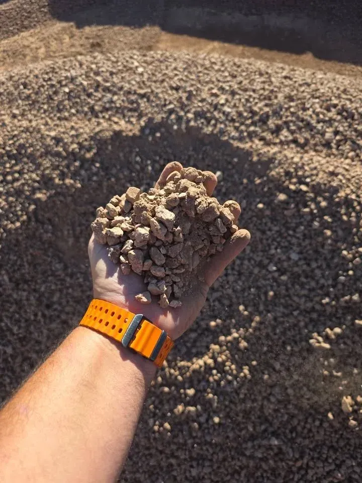Handful of gravel, held up for view. Hand has orange watch, against backdrop of gravel.