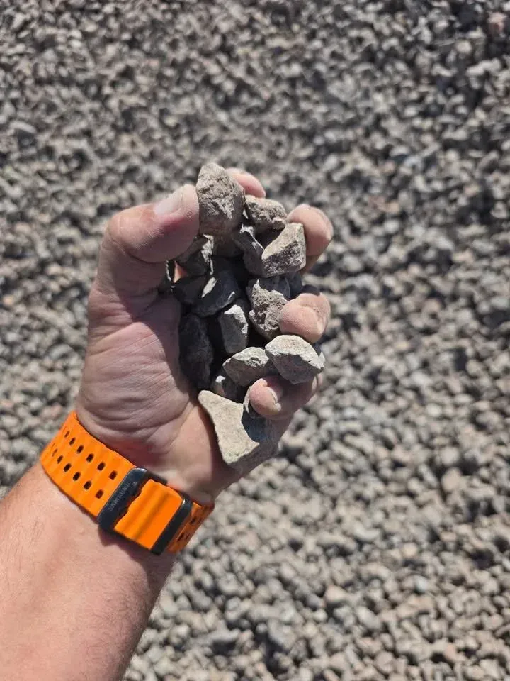 Hand holding a pile of gray gravel against a background of more gravel. The person wears an orange watch.