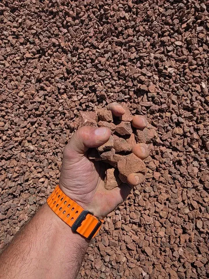 Handful of red-brown gravel. A wrist with an orange watch is visible.