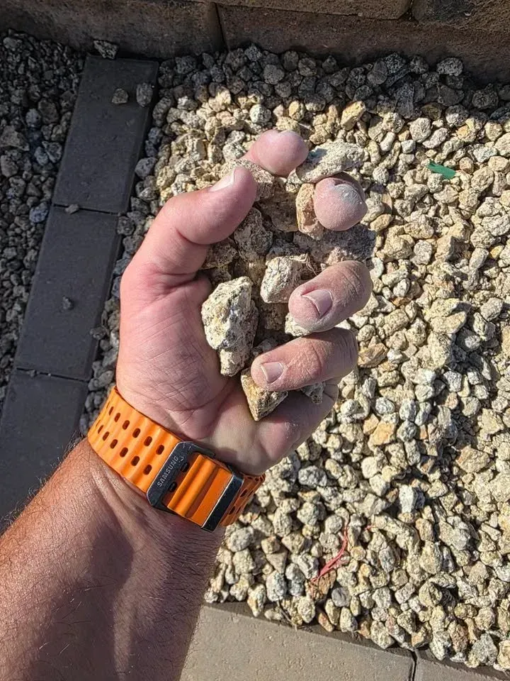 Handful of beige gravel held over a bed of similar gravel. Orange and black watch on wrist.