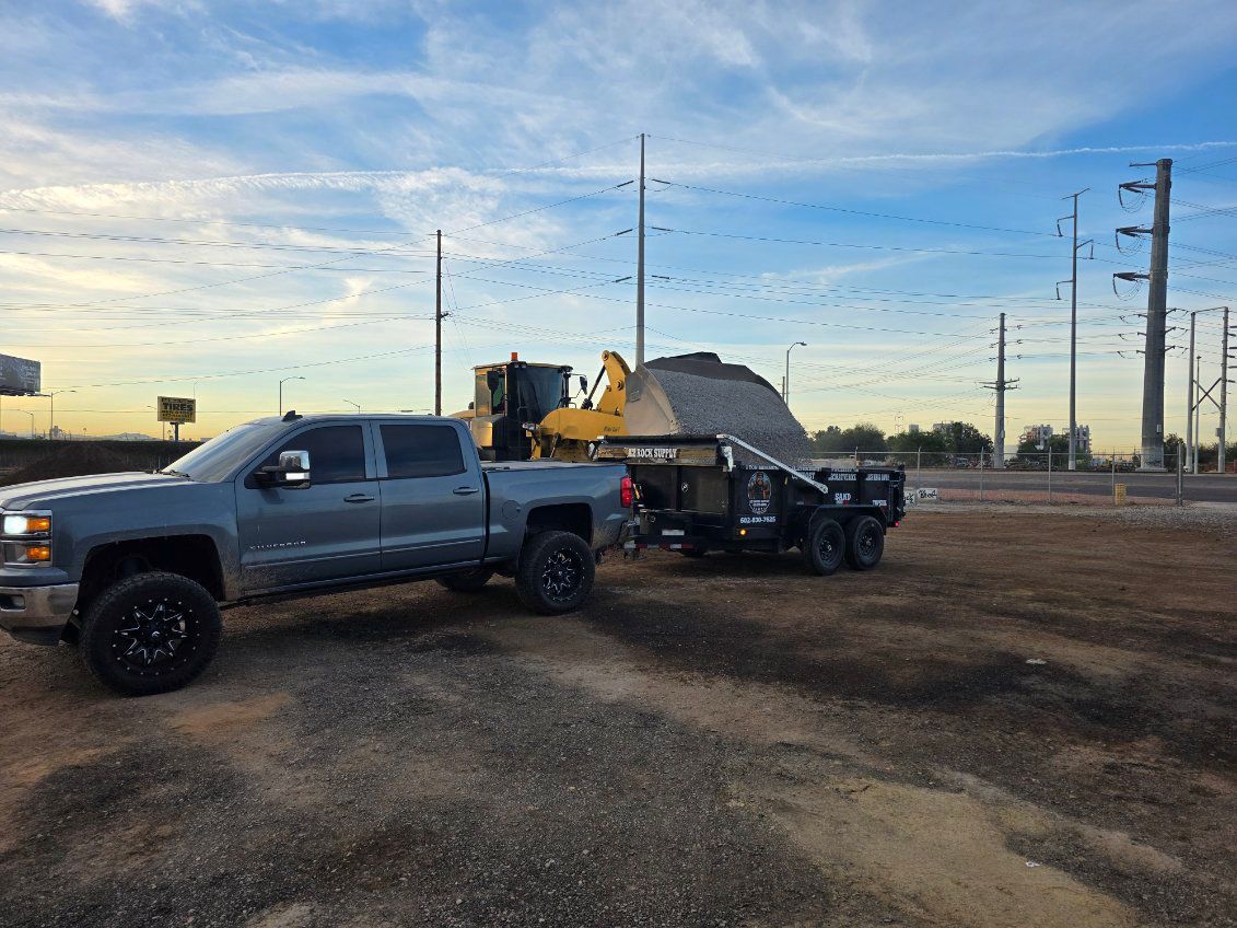 Gray pickup truck towing a trailer loaded with gravel, next to a yellow loader under a blue sky.