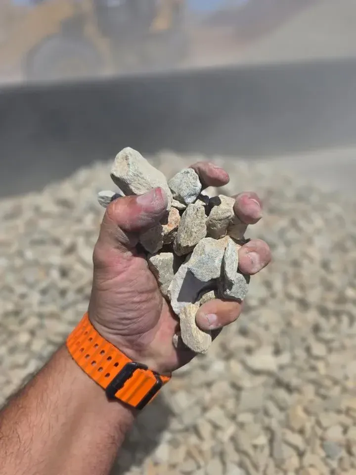 Handful of light-colored gravel. An arm with an orange watchband holds the stones with a construction site in the background.